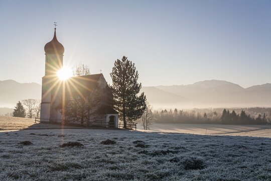 Chapel at sunrise in front of mountains, backlight, sunbeams, hoarfrost, winter, Sankt Johannisrain, Alpine foothills, Bavaria, Germany