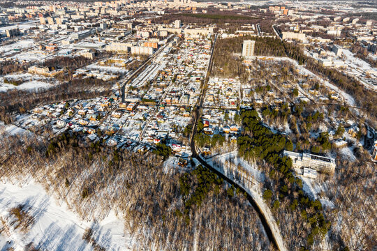 Aerial view of a suburban neighborhood nestled in a snowy winter landscape, houses peeking through bare trees and evergreen clusters under bright sunlight