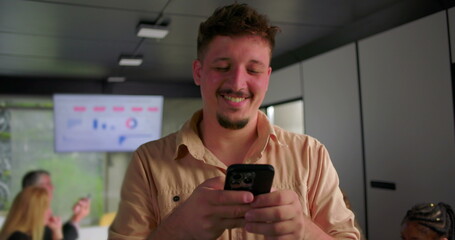 Happy young businessman uses smartphone in office, smiling while typing during team meeting with coworkers working on laptops in background