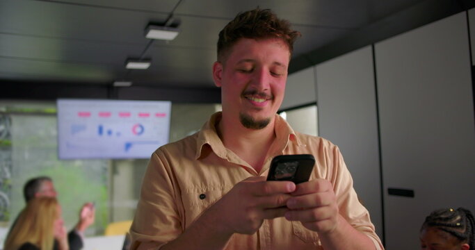 Happy young businessman uses smartphone in office, smiling while typing during team meeting with coworkers working on laptops in background - Powered by Adobe