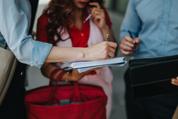 A group of professionals gathers outside as one person writes in a notebook while others hold pens and a folder, signaling teamwork, planning, and active discussion in a relaxed urban setting.
