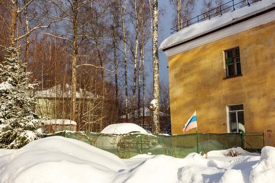 A sunlit, weathered yellow building stands against a backdrop of bare birch trees and deep winter snow, a tattered Russian flag waves near the facade, Russia