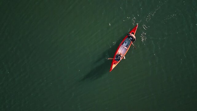 Aerial shot of a couple paddling a vibrant red canoe on calm, green waters, embodying a serene weekend activity. Their synchronized strokes reflect teamwork and connection. - Powered by Adobe