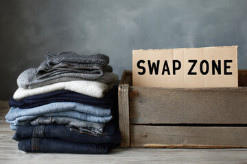 Second hand clothes swap event with folded denim jeans and sweaters stacked beside wooden crate labeled swap zone, promoting sustainable fashion and eco friendly lifestyle choices