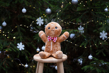 A cute gingerbread plush toy standing in front of a beautifully decorated Christmas tree, surrounded by warm holiday lights and festive ornaments. The cozy Christmas atmosphere.
