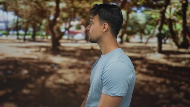 Young man in a blue shirt stands thoughtfully in a sunlit park surrounded by green trees, evoking a sense of tranquility and contemplation.