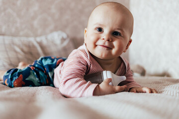 The photo shows a smiling baby lying on a soft blanket. The child is wearing a pink-and-white striped outfit and holding a baby bottle. The warm, light background gives the scene a cozy, homey feeling