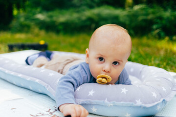 The photo shows a baby lying on a soft star-patterned cushion outdoors. The child is looking directly at the camera while sucking on a pacifier. The warm sunlight and green background create a peacefu