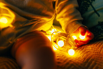 A baby is lying on pillows in warm, cozy lighting, carefully observing a glowing string of fairy lights. The soft illumination highlights the child's face, creating a magical and peaceful atmosphere. 