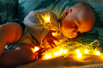 A baby is lying on pillows in warm, cozy lighting, carefully observing a glowing string of fairy lights. The soft illumination highlights the child's face, creating a magical and peaceful atmosphere. 