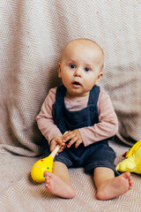 A cute baby sits on a soft chevron-patterned blanket, holding a bright yellow rattle toy. The child, dressed in a pink long-sleeve shirt and dark overalls, looks directly at the camera with a curious 