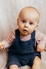 A cute baby sits on a soft chevron-patterned blanket, holding a bright yellow rattle toy. The child, dressed in a pink long-sleeve shirt and dark overalls, looks directly at the camera with a curious 