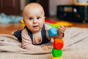 A baby lying on their stomach on a soft blanket reaches for colorful toys in a play area. Warm natural light and a cozy home environment create a bright and engaging family moment.
