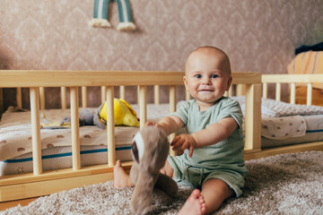 Cute baby sitting on a soft rug in a nursery next to a wooden crib. The child is holding a plush toy and looking at the camera with a gentle smile. Warm home lighting, natural tones, and a cozy atmosp