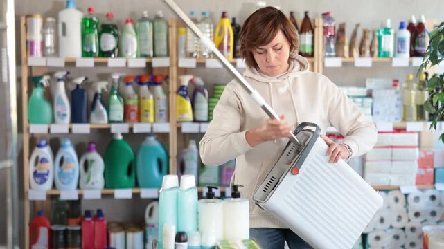 Woman housewife purchases in household chemicals store, choose mop with plastic bucket. Buyer examines product in detail, looking for consumer information on packaging