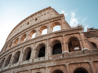 Obraz premium Low-angle photograph of the ancient Colosseum featuring its iconic stone arches, warm natural lighting, and a clear blue sky. A classic historical landmark