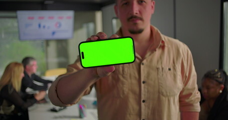Horizontal smartphone with green screen held by young man toward camera in office, team in background working near data chart on digital display