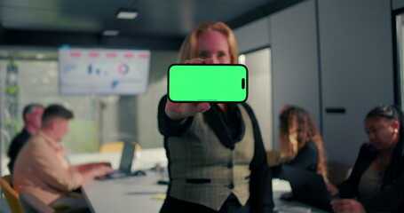 Horizontal smartphone with green screen held toward camera by businesswoman in office, coworkers working with laptops and data charts in background
