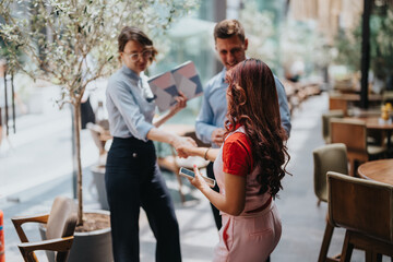 Three people in casual business attire stand near cafe seating outdoors, shaking hands while one holds a smartphone as they chat and share ideas in a relaxed city setting.