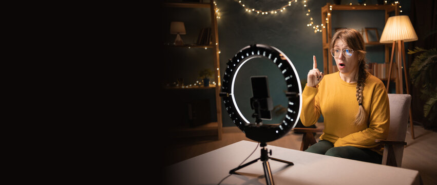 Female blogger recording video using smartphone making live selfie, social media content live video streaming making idea gesture late evening at home office. social media