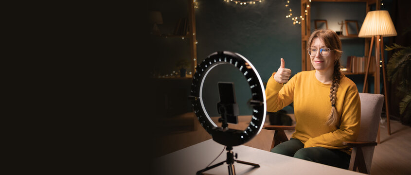 Female blogger recording video using smartphone making live selfie, social media content live video streaming making thumb-up like gesture late evening at home office. social media - Powered by Adobe
