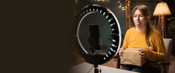 Female blogger unpacking box recording video using smartphone, ring light with tripod sticking...