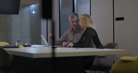 Male and female coworkers in discussion at conference table reviewing information on laptop, collaborating during late office hours in modern workplace