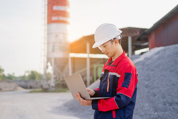 An engineer standing at industrial material storage yard checking data plans on laptop. Project management quality control. Large cement silo and pile of materials in background.