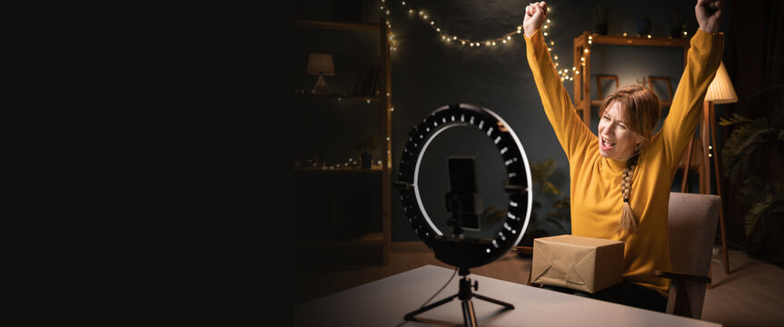Female blogger unpacking box recording video using smartphone, ring light with tripod celebrating winner late evening at home office. Concept of online shopping and social media - Powered by Adobe