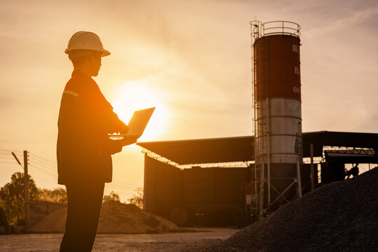 An engineer standing at industrial material storage yard checking data plans on laptop. Project management quality control. Large cement silo and pile of materials in background.