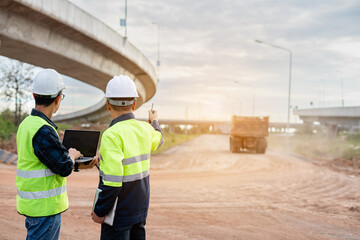 Two construction engineers, wearing hard hats and high-visibility vests, stand at a highway interchange construction site. discussing plans with his colleague who holds a walkie-talkie and tablet.