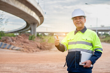Senior construction manager or engineer standing at highway construction site, holding tablet, long overpass behind successful administration of approval for large infrastructure development.