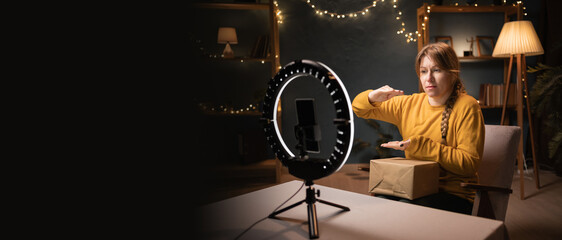 Female blogger unpacking box recording video using smartphone, ring light with tripod showing big size box gesture late evening at home office. Concept of online shopping and social media