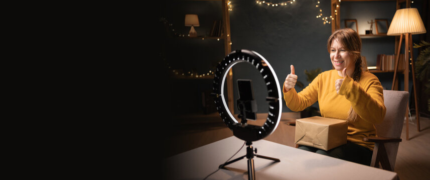 Female blogger unpacking box recording video using smartphone, ring light with tripod celebrating success with thumbs up late evening at home office. Concept of social media - Powered by Adobe