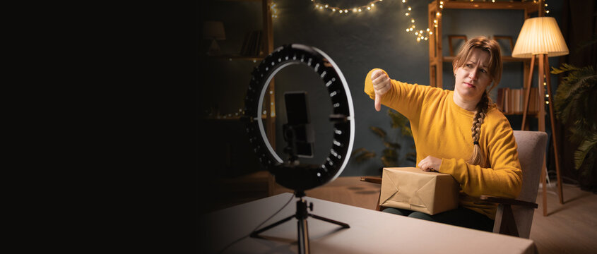 disappointed Female blogger unpacking box recording video using smartphone, ring light with tripod showing thumb down dislike gesture late evening at home office. social media