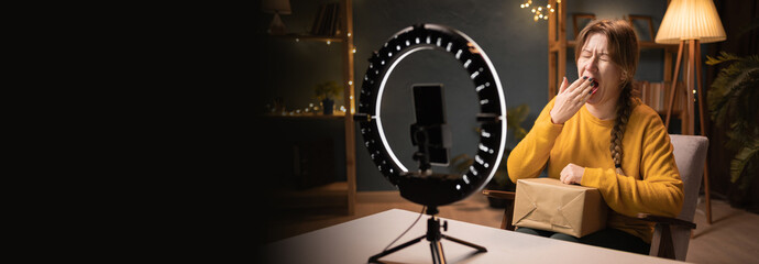 Female blogger unpacking box recording video using smartphone, ring light with tripod yawns with...