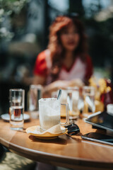 A casual outdoor cafe setting featuring a foamy latte mug on a wooden table, with a blurred figure in the background. Glass of water, napkin, and devices create a relaxed, daytime mood.