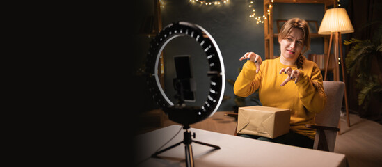 Female blogger unpacking box recording video using smartphone, ring light with tripod funny doing...