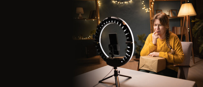 nervous Female blogger unpacking box recording video using smartphone, ring light with tripod biting her nails late evening at home office. Concept of online shopping and social media
