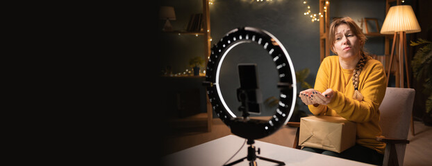 Female blogger unpacking box recording video using smartphone, ring light with tripod giving or...
