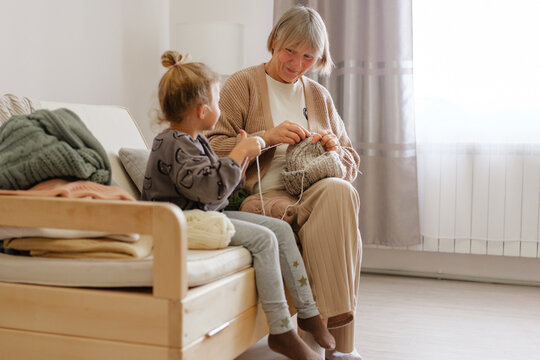 An elderly woman shares her knitting skills with a young girl in a warm and inviting living room. The afternoon light creates a peaceful atmosphere