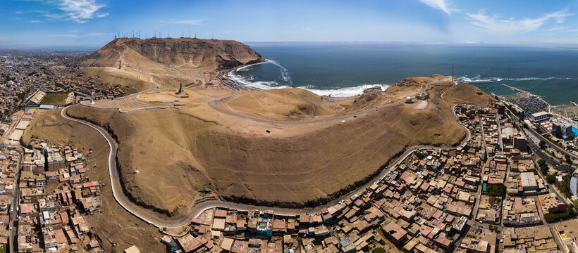 Panoramic aerial view of the Morro Solar and La Chira hills in Chorrillos, Lima, featuring communication antennas, winding roads, and the Pacific Ocean coastline.