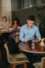 A focused man in a light blue shirt sits at a round cafe table, using a tablet with a stylus. A phone and glass of water sit nearby, with colleagues blurred in the background.