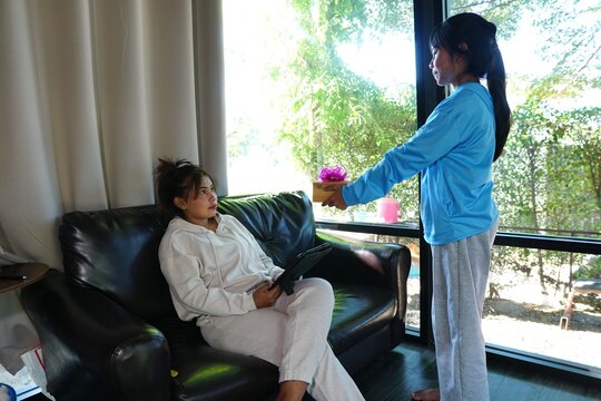 The daughter presents a small gift box with flowers to her mother, who is seated on the couch.
