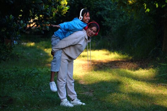 The mother is giving her daughter a piggyback ride while both are smiling and posing like they are flying in the park.