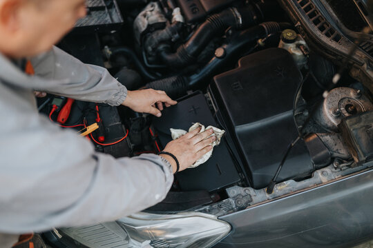 A technician wipes the car battery area under the hood, surrounded by tools and cables. A focused moment of maintenance and cleaning in a vehicle workshop.