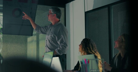Male presenter pointing to chart on screen while team listens during modern office meeting with digital data visualization