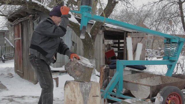 A man works on a homemade wood splitter in a snowy yard. A man splits a wooden log with mechanical wood splitter, Preparing firewood for winter using mechanical press.