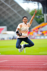 A young male photographer jumping joyfully on an outdoor stadium track while holding a professional telephoto camera