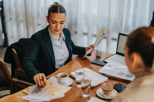 Two young coworkers collaborate over charts and papers at a bright office table, with coffee and a laptop nearby, conveying focus and teamwork in a professional setting. - Powered by Adobe